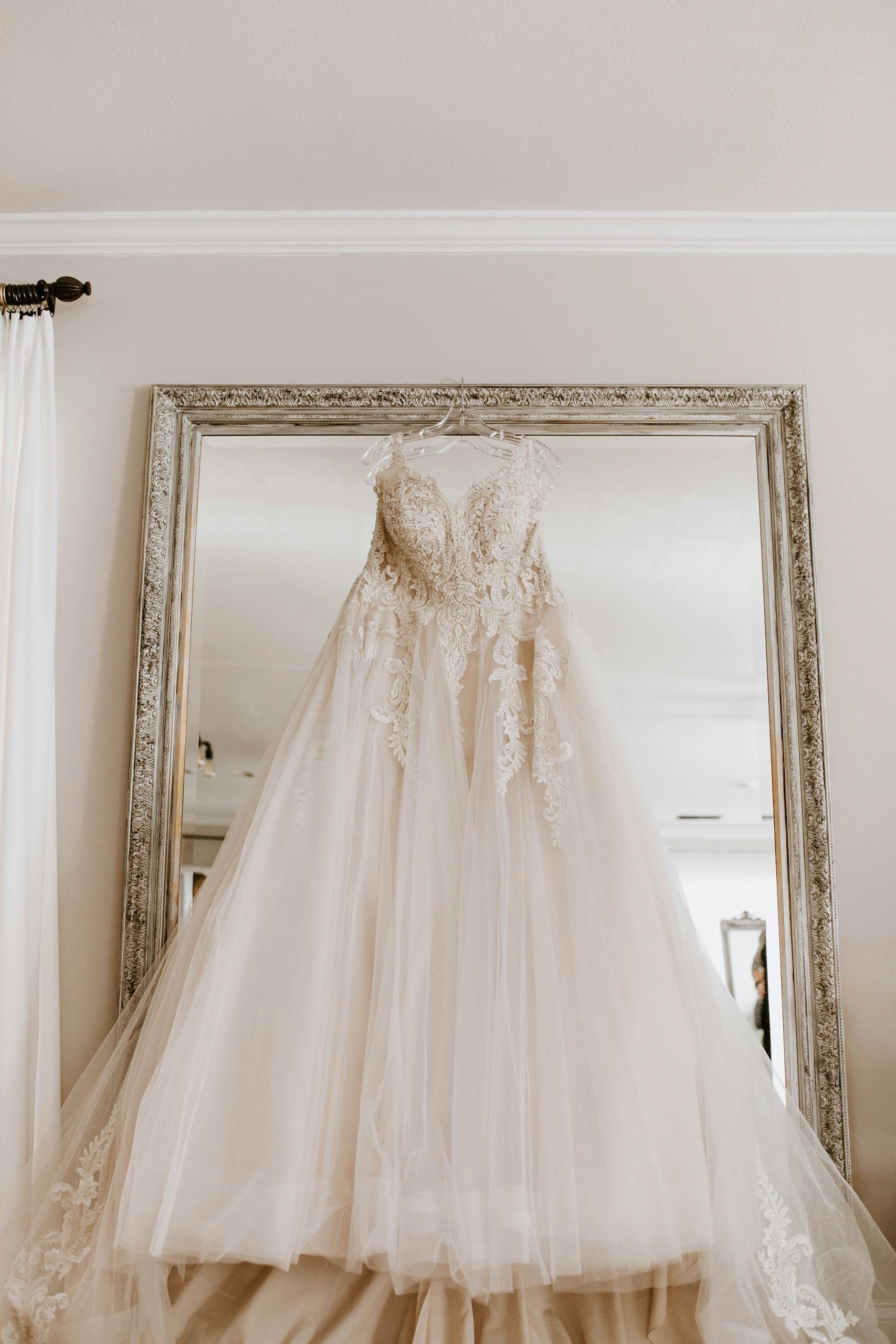 Low angle of elegant white bridal dress hanging on big framed mirror in light room before wedding ceremony