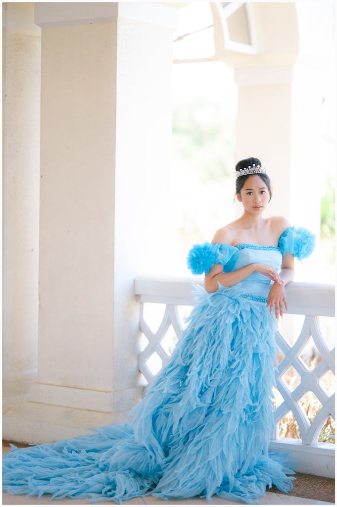 An elegant woman in a blue gown with a crown, posing against a column outdoors.
